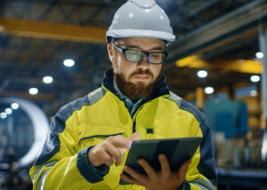 Industrial Engineer in Hard Hat Wearing Safety Jacket Uses Touchscreen Tablet Computer. He Works at the Heavy Industry Manufacturing Factory.